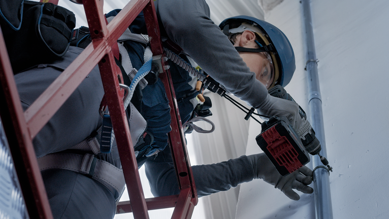 Eine Person mit Sicherheitsausrüstung bohrt mit einem Elektrowerkzeug von einem Gerüst aus in eine Wand.