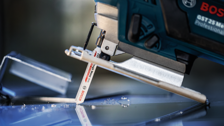 Eine Stichsäge schneidet durch eine Glasscheibe, in deren Nähe verstreute Glassplitter liegen.