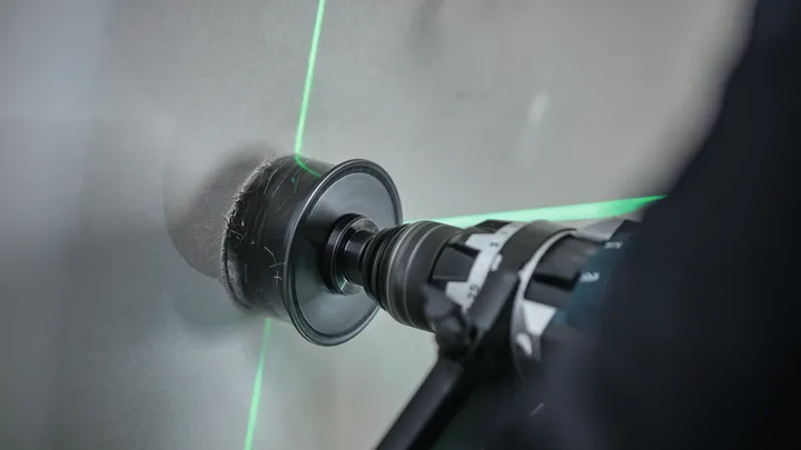 Eine Person verwendet eine Bohrmaschine mit Lochsäge, um Fliesen zu schneiden, wobei sie sich an grünen Laserlinien orientiert.