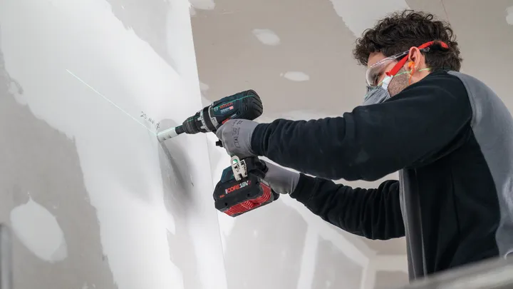 Eine Person mit Sicherheitsausrüstung bohrt entlang einer grünen Laserführung in eine Trockenbauwand.