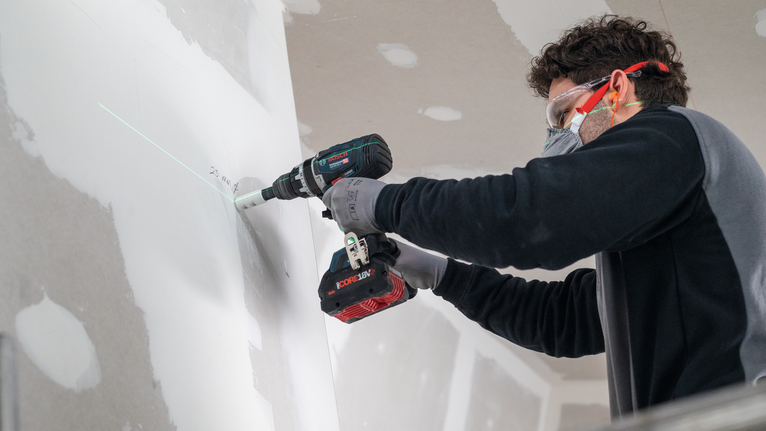 Eine Person mit Sicherheitsausrüstung bohrt entlang einer grünen Laserführung in eine Trockenbauwand.