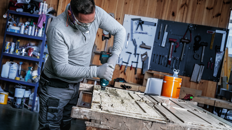 Eine Person, die Schutzausrüstung trägt, bohrt auf einem Werkstatttisch in eine Holztür.
