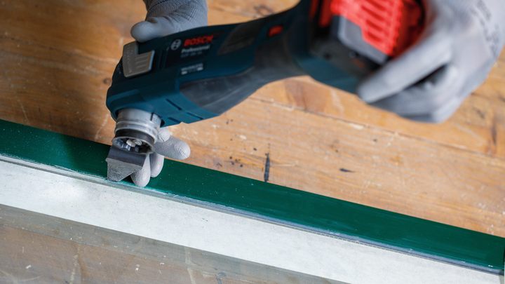 Eine Person mit Schutzausrüstung schleift mit einem Akku-Multifunktionswerkzeug einen lackierten Fensterrahmen.