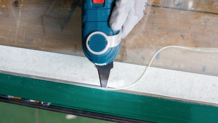 Eine Person mit Sicherheitsausrüstung entfernt mit einem Elektrowerkzeug Dichtungsmittel von einem Fensterrahmen.