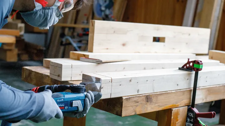 Eine Person mit Sicherheitsausrüstung verwendet ein Elektrowerkzeug, um Holzbalken zu schneiden.