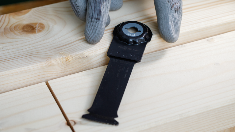 Eine Person mit Sicherheitsausrüstung hält ein Multitool-Sägeblatt auf einem Holzbrett.