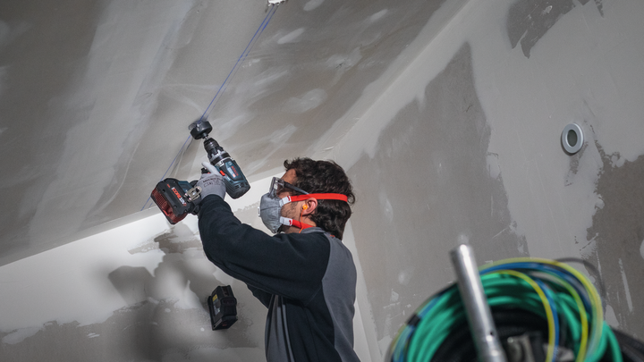 Eine Person mit Sicherheitsausrüstung bohrt in eine Decke auf einer Baustelle.