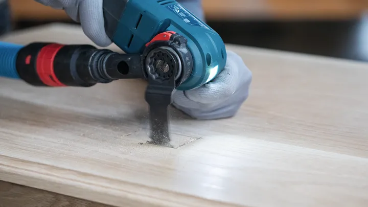 Eine Person mit Sicherheitsausrüstung schneidet mit einem Elektrowerkzeug ein rechteckiges Loch in Holz.