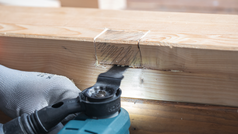 Eine Person mit Sicherheitsausrüstung verwendet ein Elektrowerkzeug, um einen Holzbalken zu schneiden.