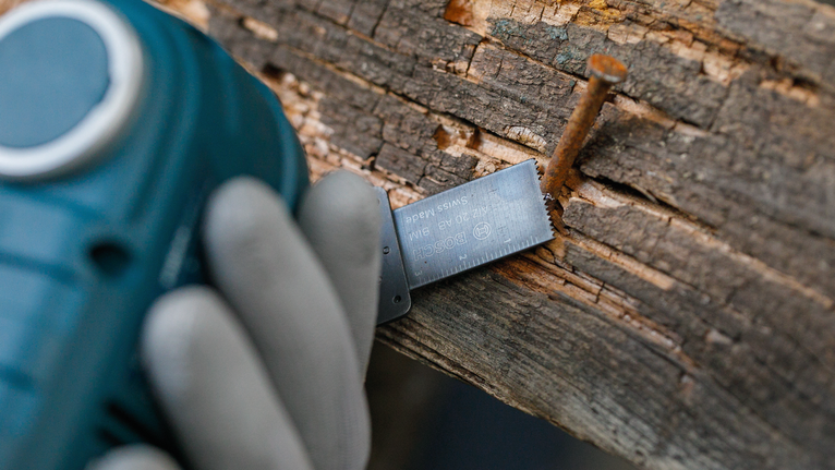 Eine Person mit Sicherheitsausrüstung schneidet mit einem Elektrowerkzeug Holz in der Nähe eines rostigen Nagels.