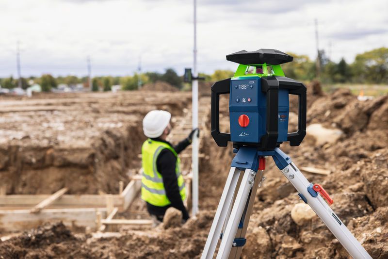 Laser-Nivelliergerät auf Stativ auf einer Baustelle mit einer Person in Schutzausrüstung.