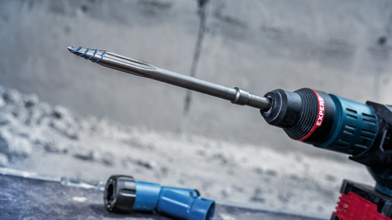 Ein Bohrhammer mit langem Bohrmeißel und blauem Werkzeugteil auf einer Betonoberfläche.