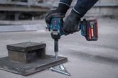 Eine Person, die Schutzausrüstung trägt, benutzt einen Akku-Schlagschrauber, um einen Bolzen in Beton zu treiben.