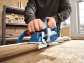 Eine Person benutzt in einer Werkstatt einen Akku-Hobel an einem Holzbrett.