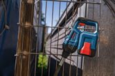 Ein blau-rotes Elektrowerkzeug lehnt im Freien an einem Metalldrahtzaun.