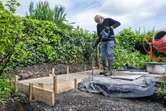 Eine Person, die Schutzausrüstung trägt, ebnet mit einem Elektrowerkzeug frischen Beton in einem Garten.