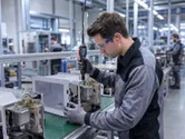 Eine Person, die Schutzausrüstung trägt, montiert an einer Produktionslinie ein Gerät mit einem Akkuschrauber.