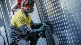 Eine Person, die Schutzausrüstung trägt, benutzt ein Elektrowerkzeug an einer metallischen Wandplatte.