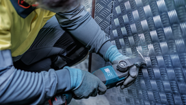 Eine Person in Schutzausrüstung schneidet mit einem Elektrowerkzeug ein gemustertes Metallblech.