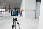 Ein auf einem Stativ montiertes Laser-Nivelliergerät projiziert eine Linie, während eine mit Schutzausrüstung ausgestattete Person eine Wand markiert.