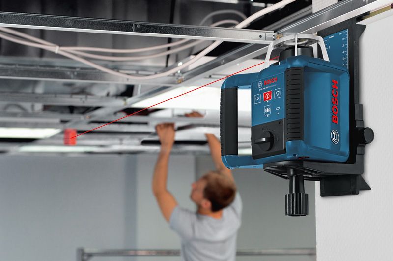 Ein an der Wand montiertes Laser-Nivelliergerät projiziert eine rote Linie, während eine Person an der Decke arbeitet.