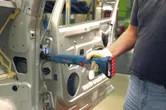 Une personne portant un équipement de sécurité utilise un outil sans fil pour fixer une charnière à l'intérieur d'une portière de voiture.