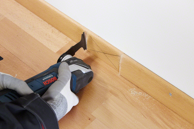 Une personne portant un équipement de sécurité coupe une plinthe en bois avec un outil multifonction.