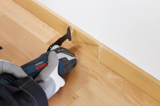 Une personne portant un équipement de sécurité coupe une plinthe en bois avec un outil multifonction.