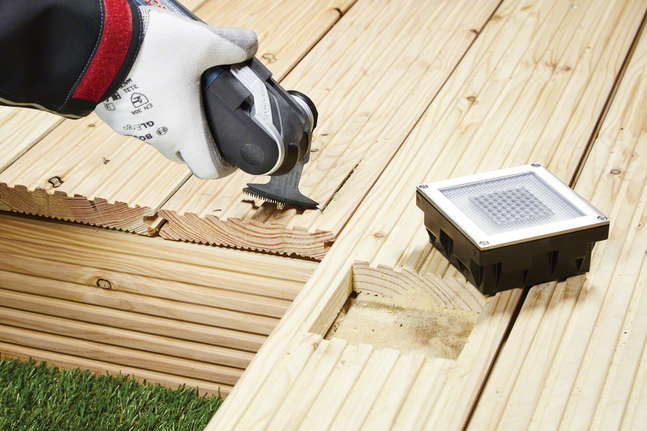 Une personne portant un équipement de sécurité coupe une terrasse en bois pour y installer un luminaire.