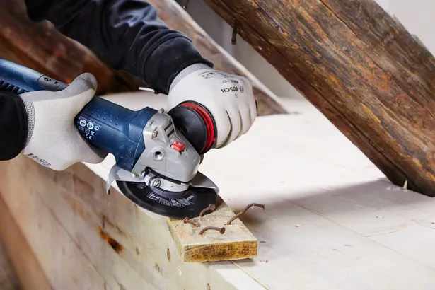Un travailleur portant un équipement de sécurité utilise une meuleuse d'angle pour couper des clous dans une planche de bois.