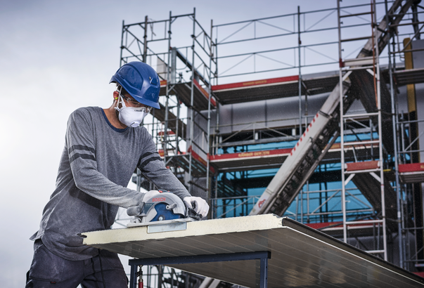Une personne portant un équipement de sécurité coupe un grand panneau avec une scie circulaire sur un chantier de construction.