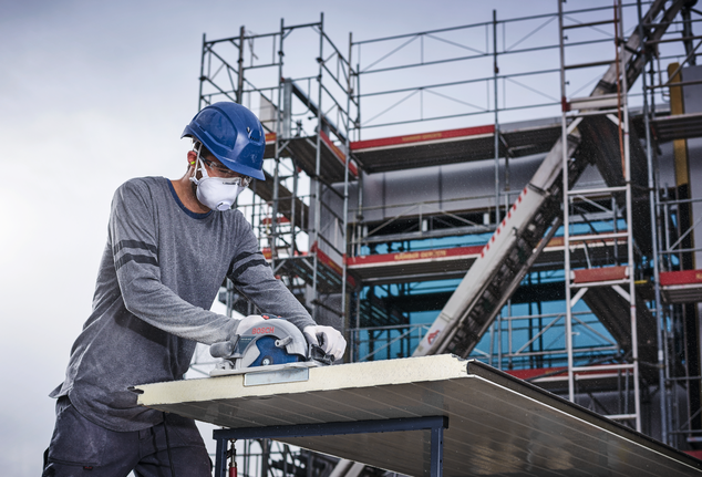 Une personne portant un équipement de sécurité coupe un grand panneau avec une scie circulaire sur un chantier de construction.