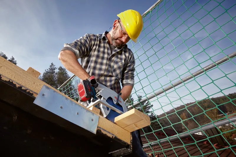Une personne portant un équipement de sécurité utilise une scie circulaire sans fil pour couper du bois sur un chantier de construction.
