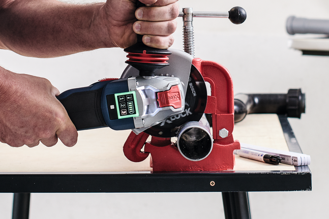 Une personne utilise une meuleuse d'angle pour couper un tuyau métallique maintenu dans un étau rouge.