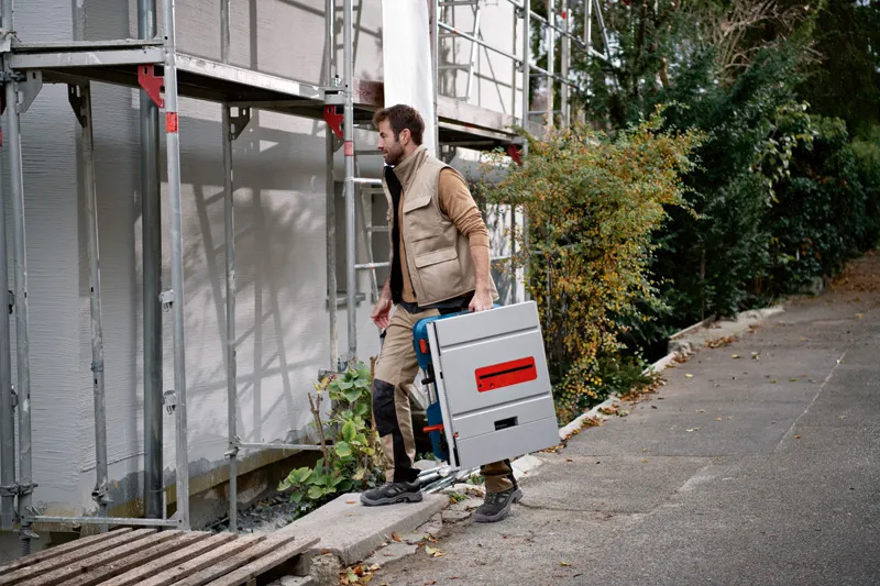 Une personne portant un équipement de sécurité transporte une scie à table sans fil sur un chantier de construction.