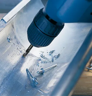 Perceuse sans fil créant un trou dans un canal métallique à proximité de rivets métalliques.