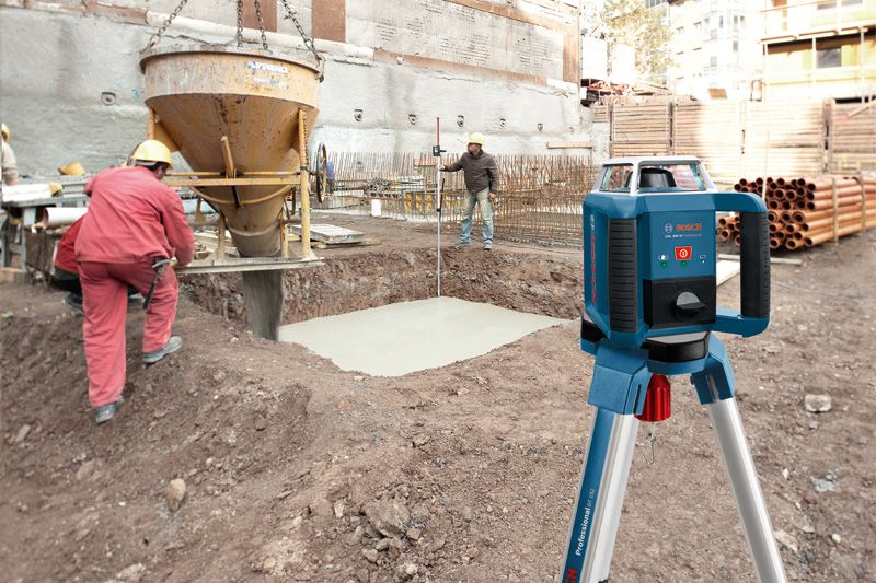 Outil de nivellement laser sur trépied sur un chantier de construction, des ouvriers portant un équipement de sécurité coulent du béton.