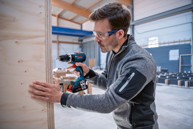 Une personne portant un équipement de sécurité perce une planche de bois à l'intérieur d'un atelier.