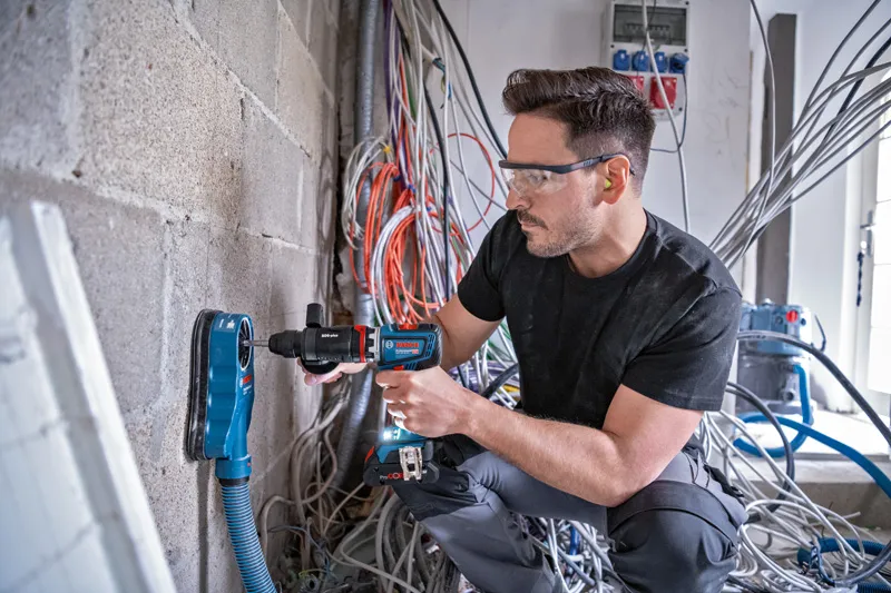 Une personne portant un équipement de sécurité perce un mur en béton avec extraction de poussière.