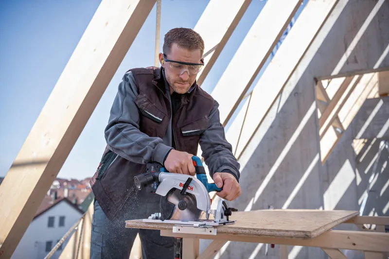 Une personne portant un équipement de sécurité utilise une scie circulaire pour couper du contreplaqué sur un chantier de construction.