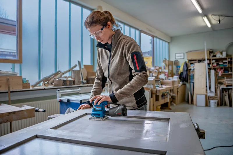 Une personne portant un équipement de sécurité ponce une porte en bois dans un atelier.