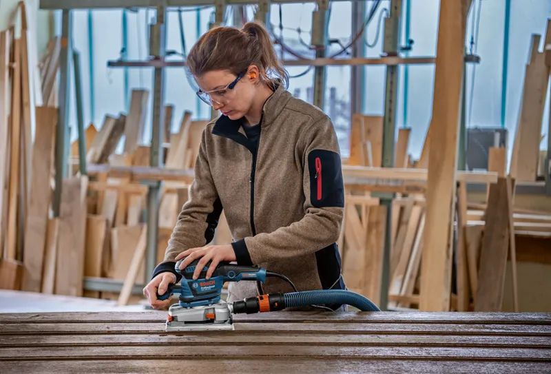Une personne portant un équipement de sécurité utilise une ponceuse orbitale sur des planches de bois dans un atelier.