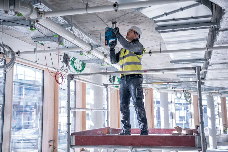 Une personne portant un équipement de sécurité perce un plafond en béton sur un chantier de construction.