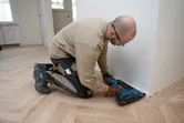Une personne portant un équipement de sécurité installe des garnitures avec une cloueuse à bois sans fil sur un plancher en bois dur.