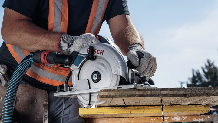Une personne portant un équipement de sécurité utilise une scie circulaire pour couper du bois à l’extérieur.