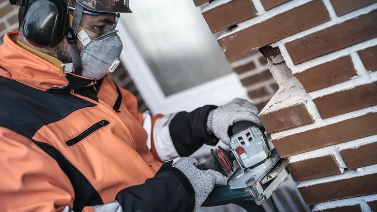 Un ouvrier portant un équipement de sécurité découpe des rainures dans un mur de briques à l'aide d'un outil électrique.