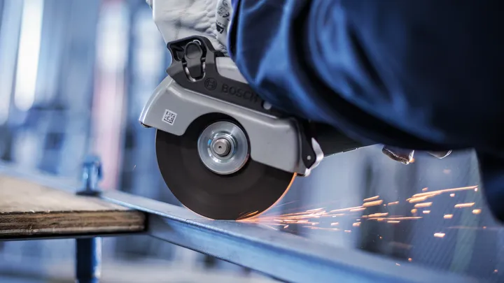 Un travailleur portant un équipement de sécurité utilise une meuleuse d'angle pour couper le métal, produisant des étincelles.