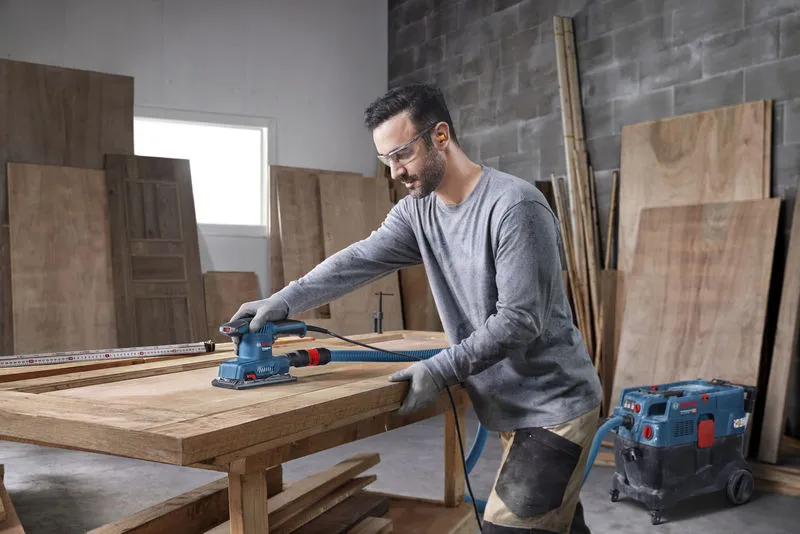 Une personne portant un équipement de sécurité ponce un plateau de table en bois avec une ponceuse orbitale.