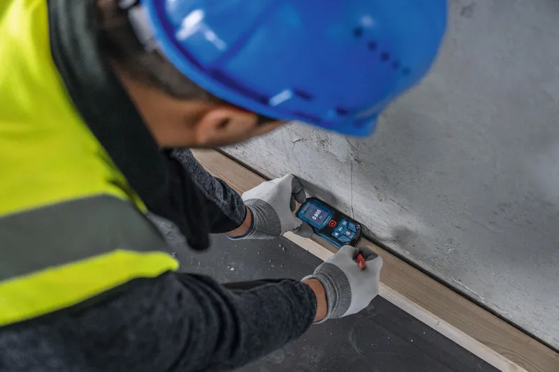 Une personne portant un équipement de sécurité mesure la distance du mur à l'aide d'un télémètre laser.