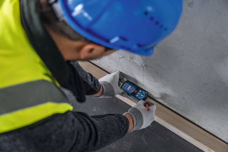 Une personne portant un équipement de sécurité mesure la distance du mur à l'aide d'un télémètre laser.